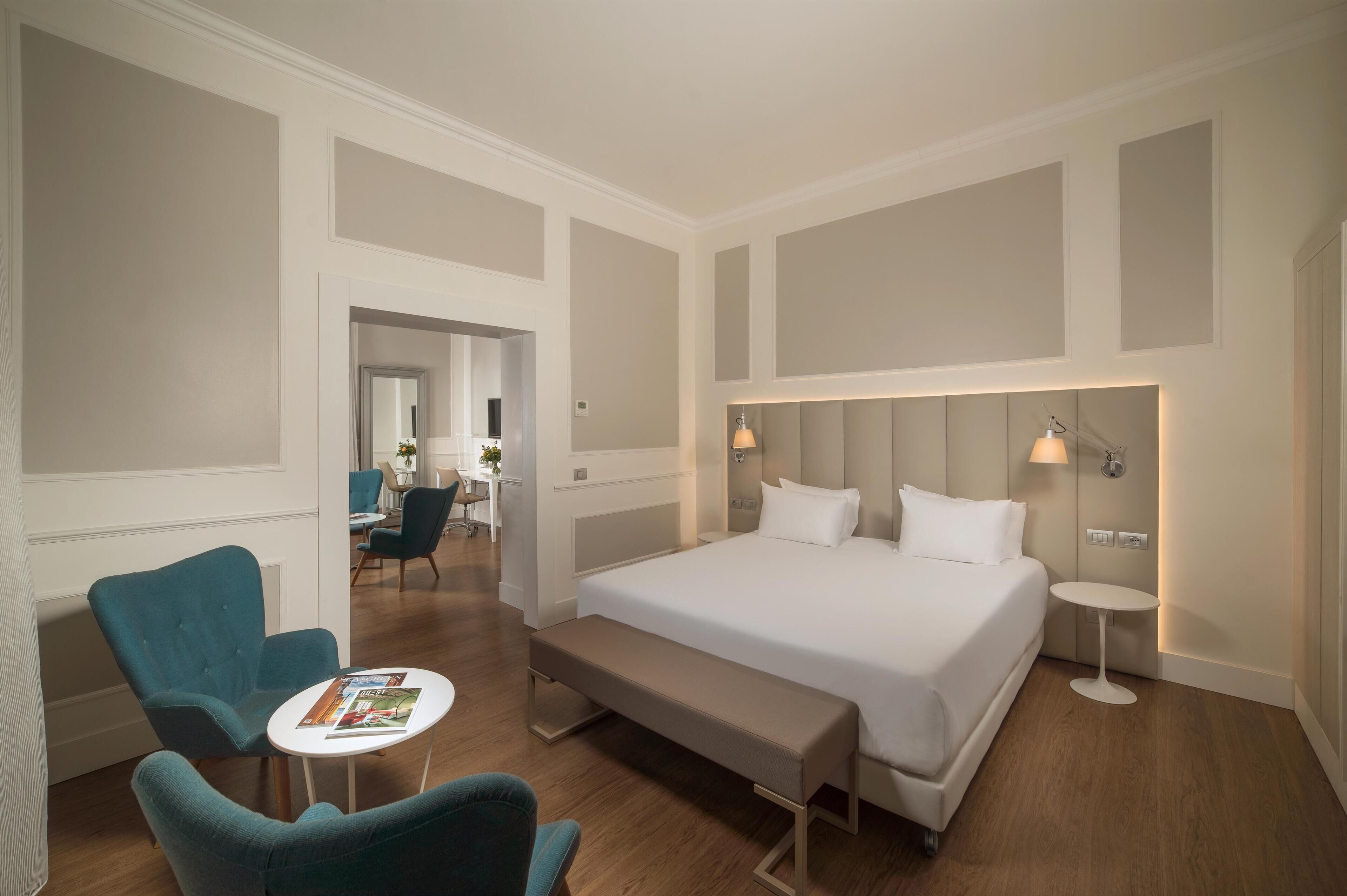Une chambre d’hôtel avec un grand lit, une banquette, deux fauteuils, une petite table et un bureau avec une chaise.