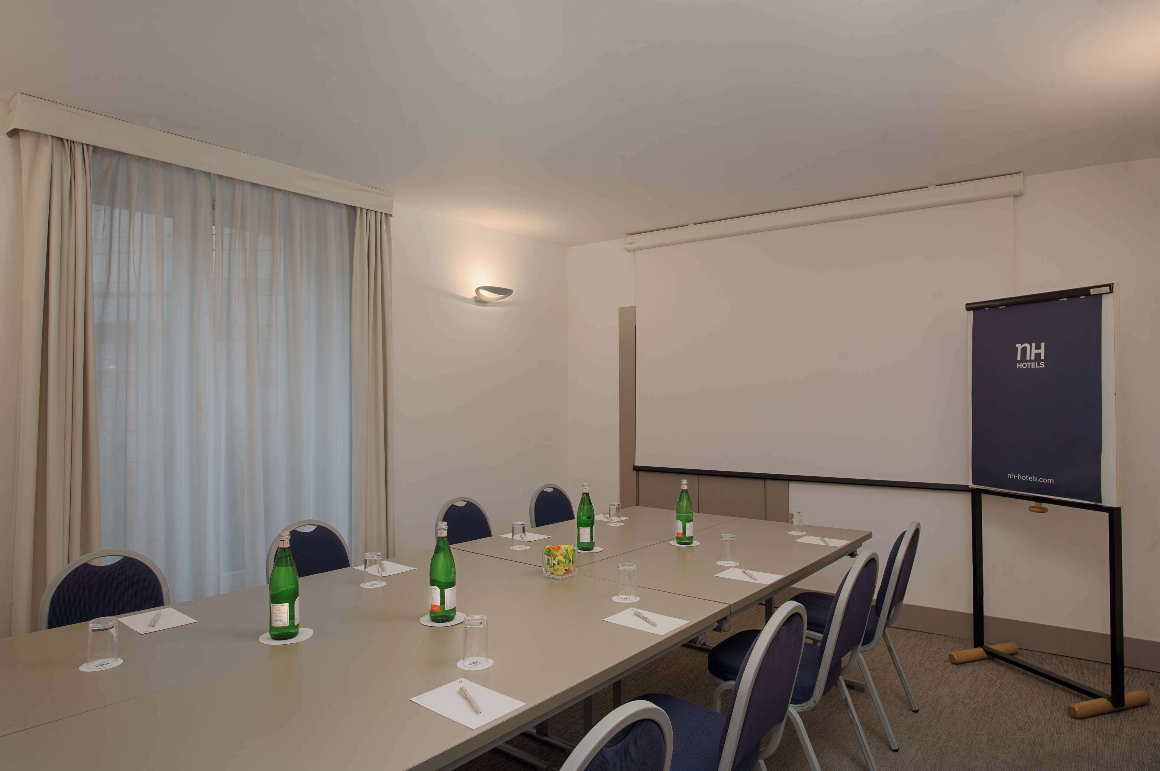 Une salle de conférence avec une longue table, des chaises, un écran de projection et une fenêtre avec des rideaux.