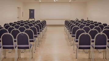 Sala de conferências com fileiras de cadeiras voltadas para uma tela de projetor, um pódio e uma porta.