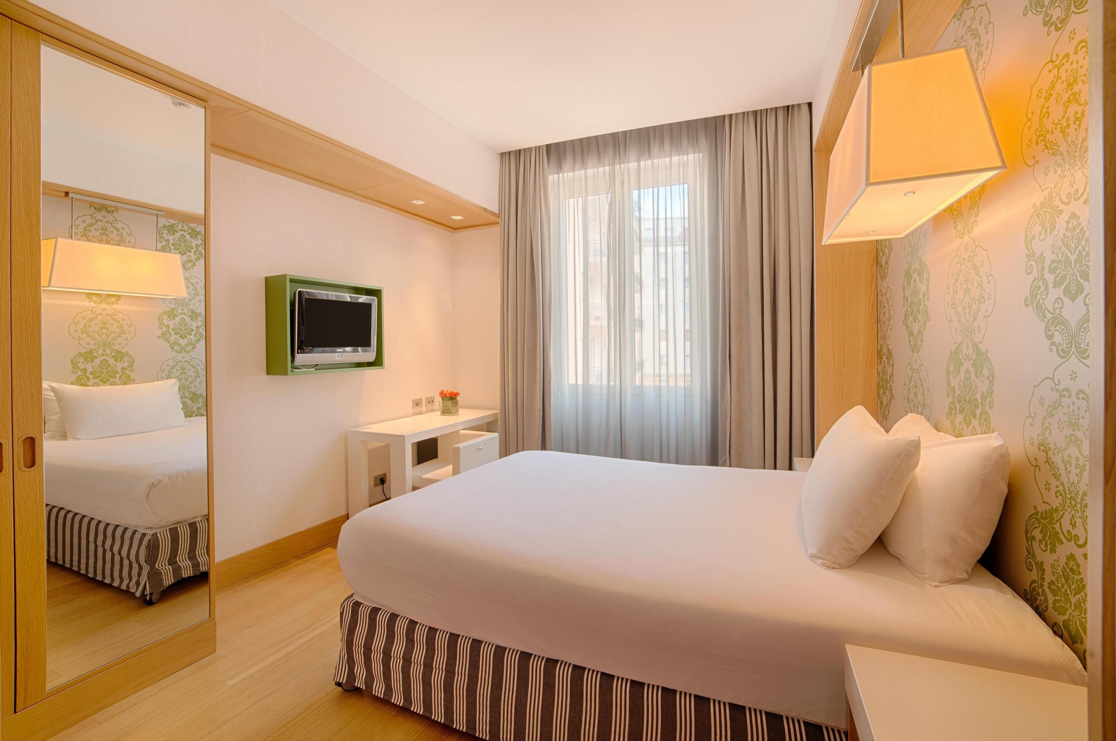 Ein Hotelzimmer mit einem großen Bett, einem Fernseher, einem Schreibtisch mit Lampe und einem Fenster mit Vorhängen.