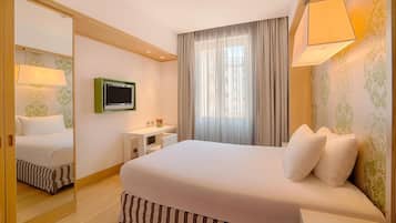 Ein Hotelzimmer mit einem großen Bett, einem Fernseher, einem Schreibtisch mit Lampe und einem Fenster mit Vorhängen.
