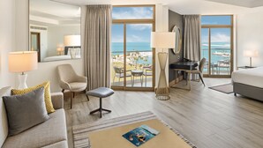 Habitación de hotel con un ventanal que ofrece vistas al mar y un balcón. Incluye una cama, un escritorio, una silla y un sofá.