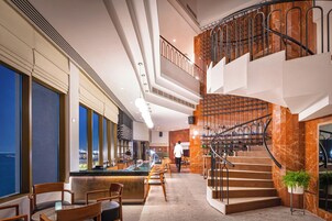 A modern hotel lobby with a bar area, seating, a staircase, and a view of the city.