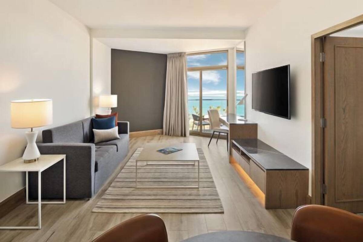 A modern hotel room with a sofa, a dining table, a TV, and a view of the ocean.