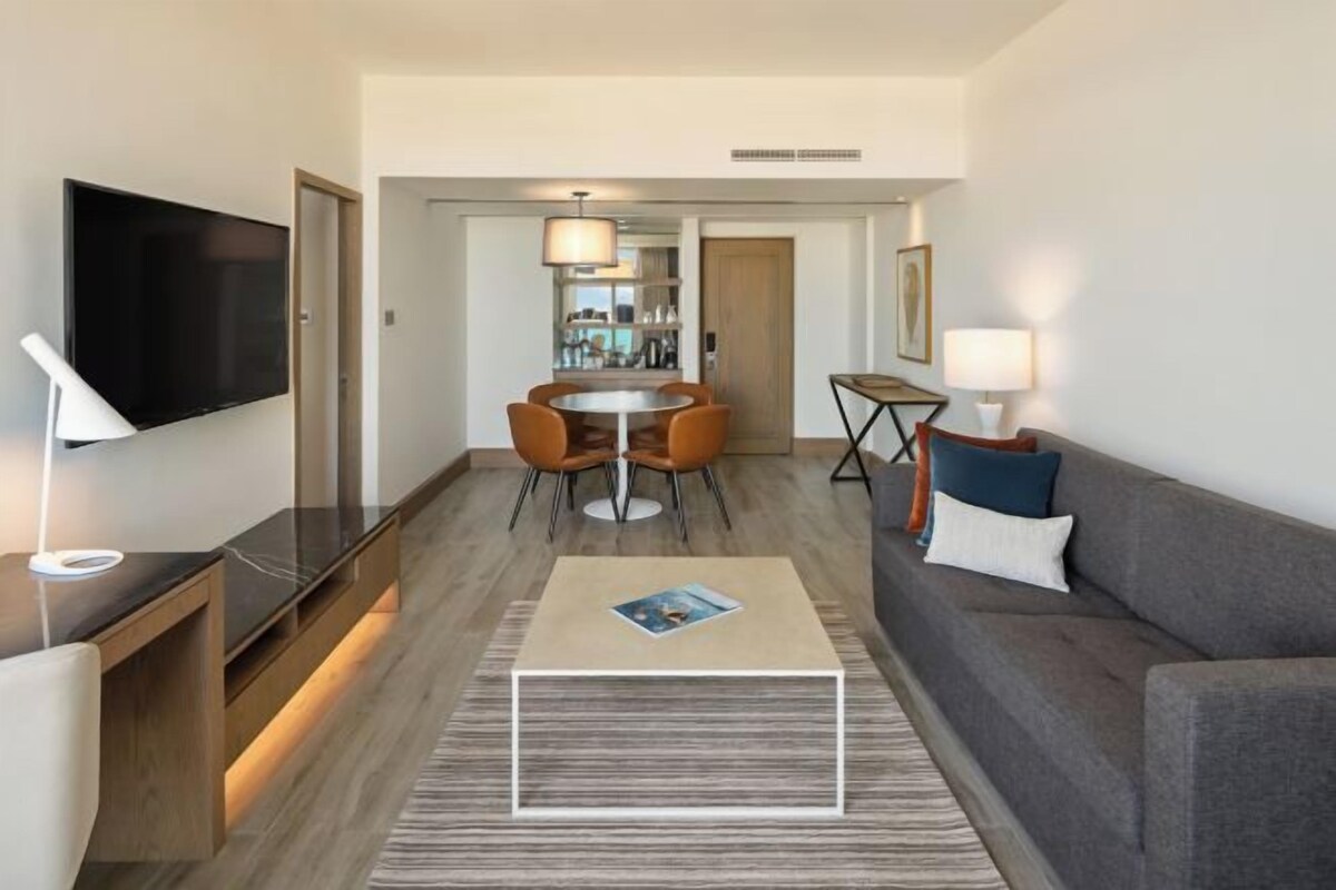 Una habitación de hotel moderna con una televisión de pantalla plana, una zona de comedor con una mesa redonda y sillas, un sofá con cojines y una mesa de centro.