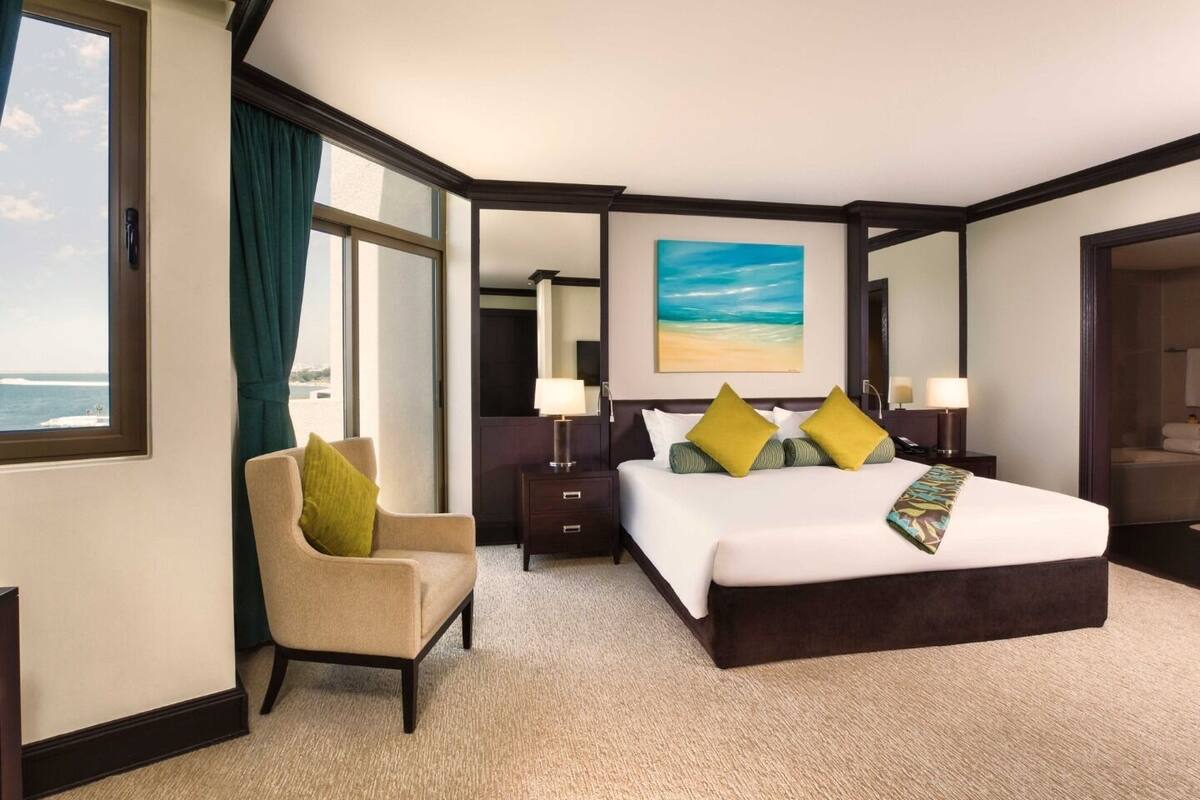 A hotel room with a large bed, a chair, a desk, and a painting of a beach.