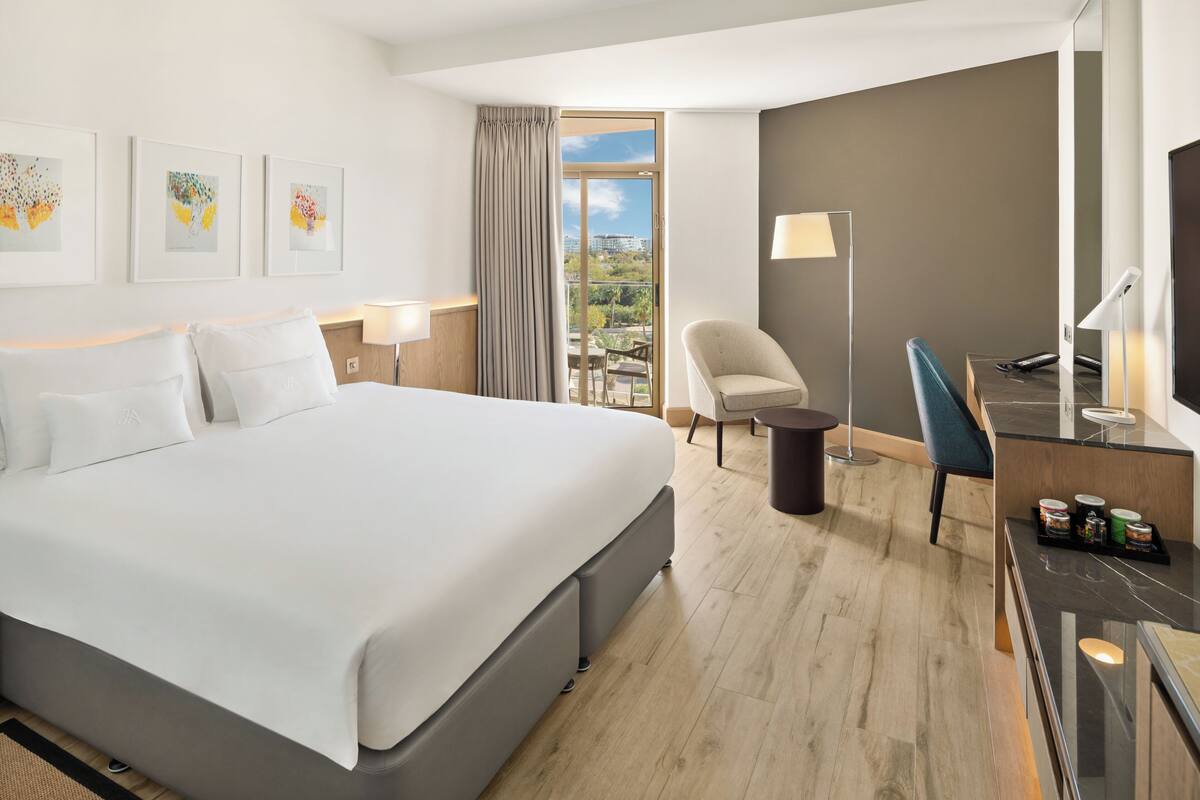 A hotel room with a large bed, a desk with a computer, a chair, a lamp, and a view of the city through a window.