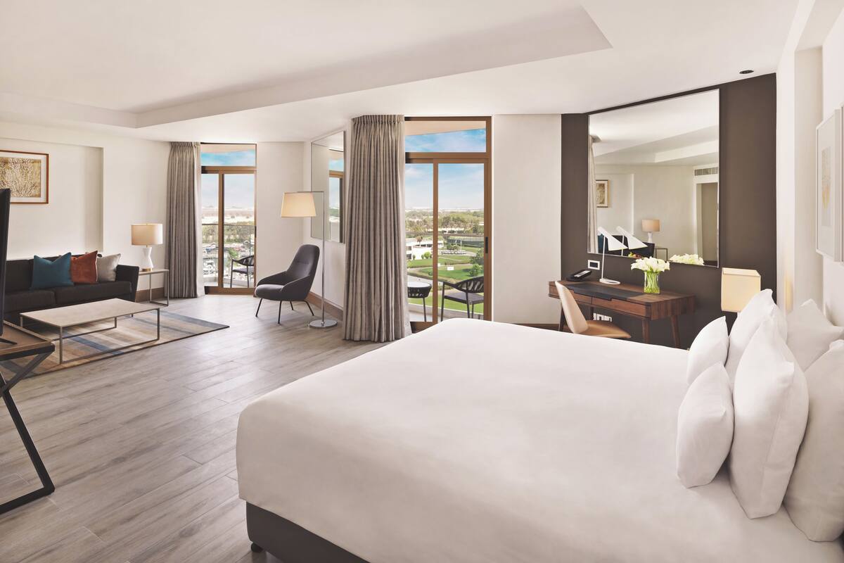 A hotel room with a large bed, a sofa, a desk, and a chair. There is a large window with curtains, a lamp, and a view of the city.