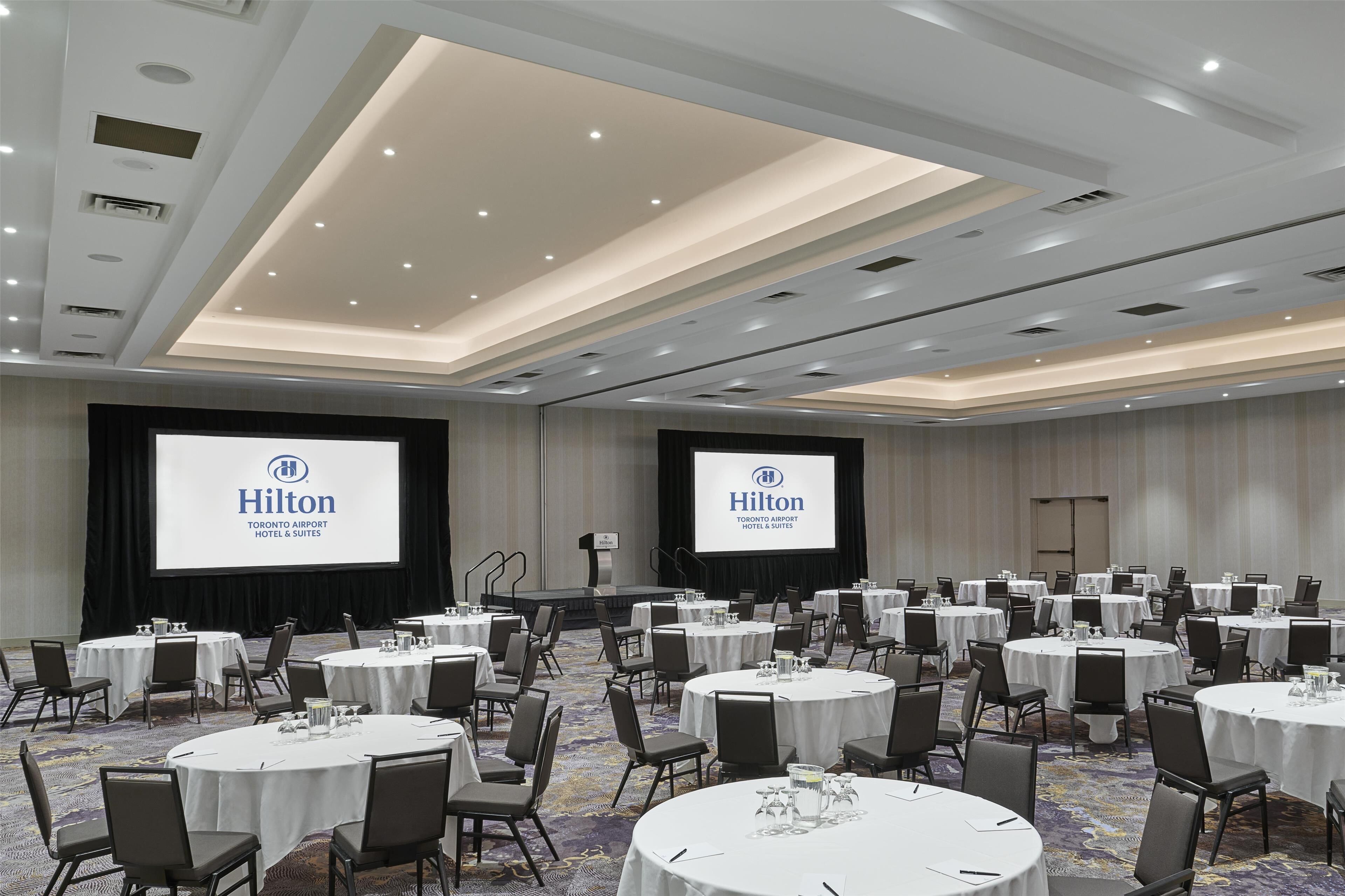 A conference room with multiple round tables, chairs, and a large screen displaying the Hilton logo.