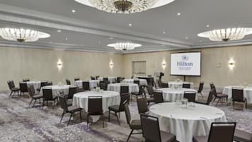 A conference room with multiple tables and chairs, a large screen displaying the Hilton logo, and elegant chandeliers.