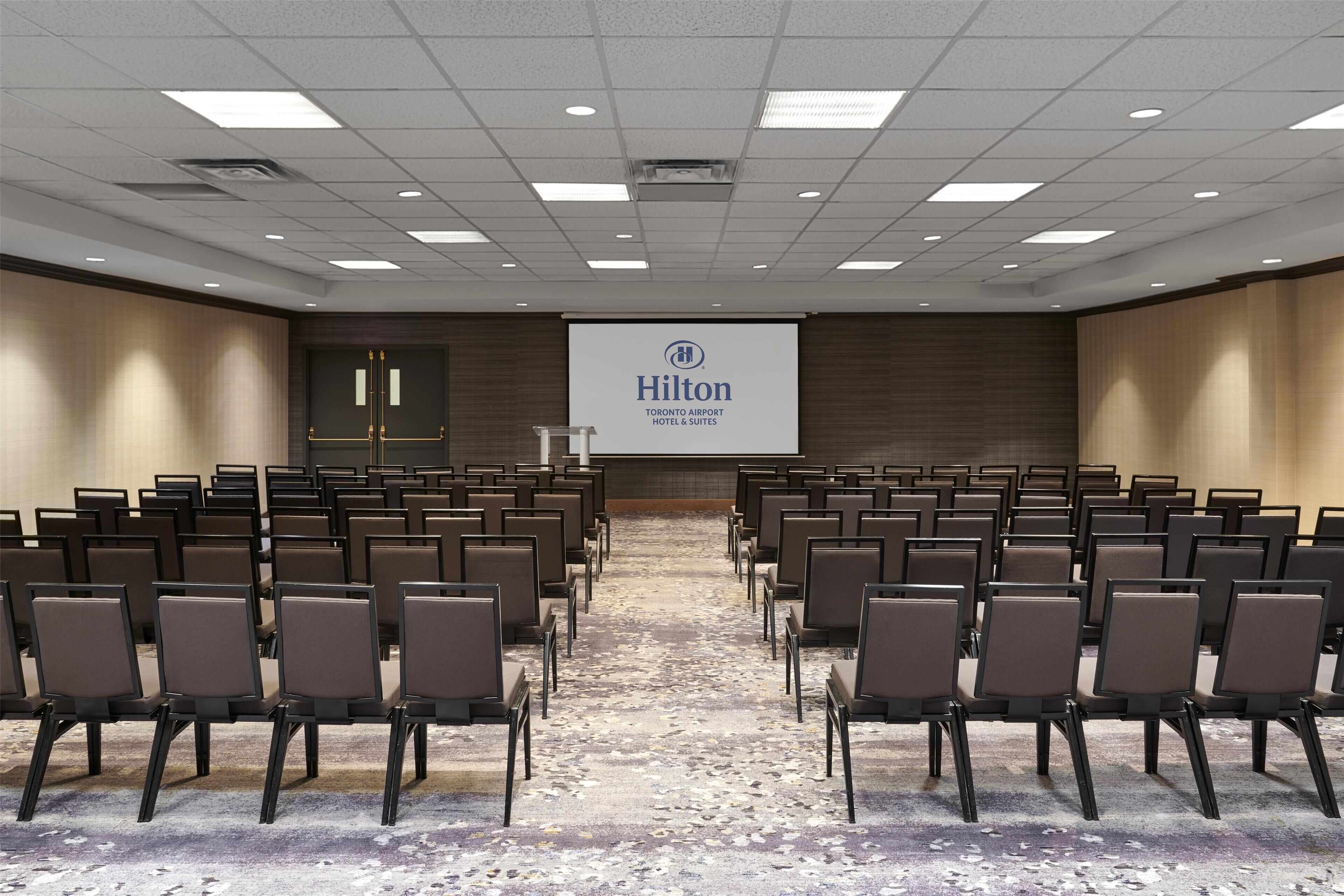 A conference room with rows of chairs, a podium, and a screen displaying the Hilton logo.