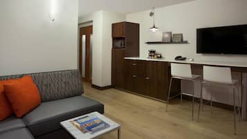 A modern living room with a sofa, coffee table, and a flat-screen TV. There is a kitchen area with a microwave, coffee maker, and a TV on a shelf.