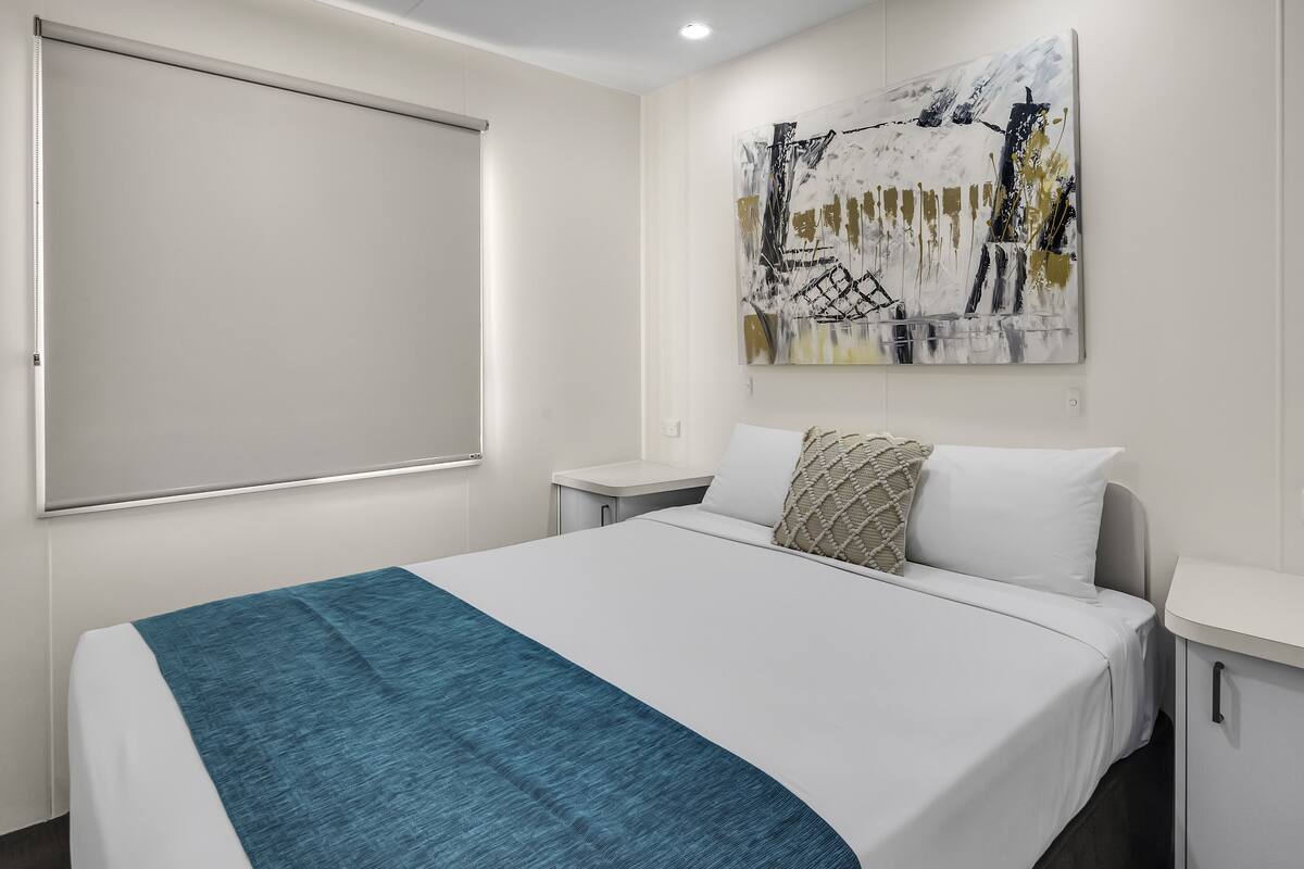 A hotel room with a bed, a bedside table, a wall-mounted artwork, and a window with a blind.