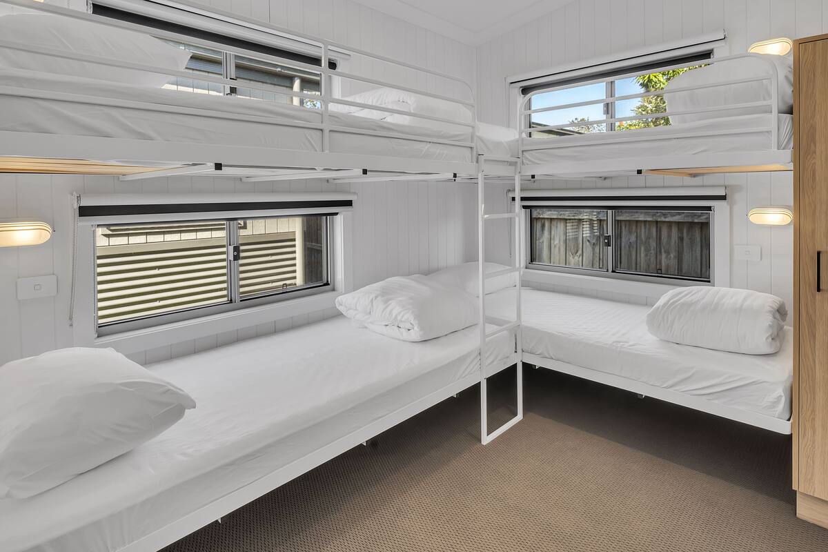 A bunk bed room with two windows, each with blinds, and a small bedside table with a lamp.