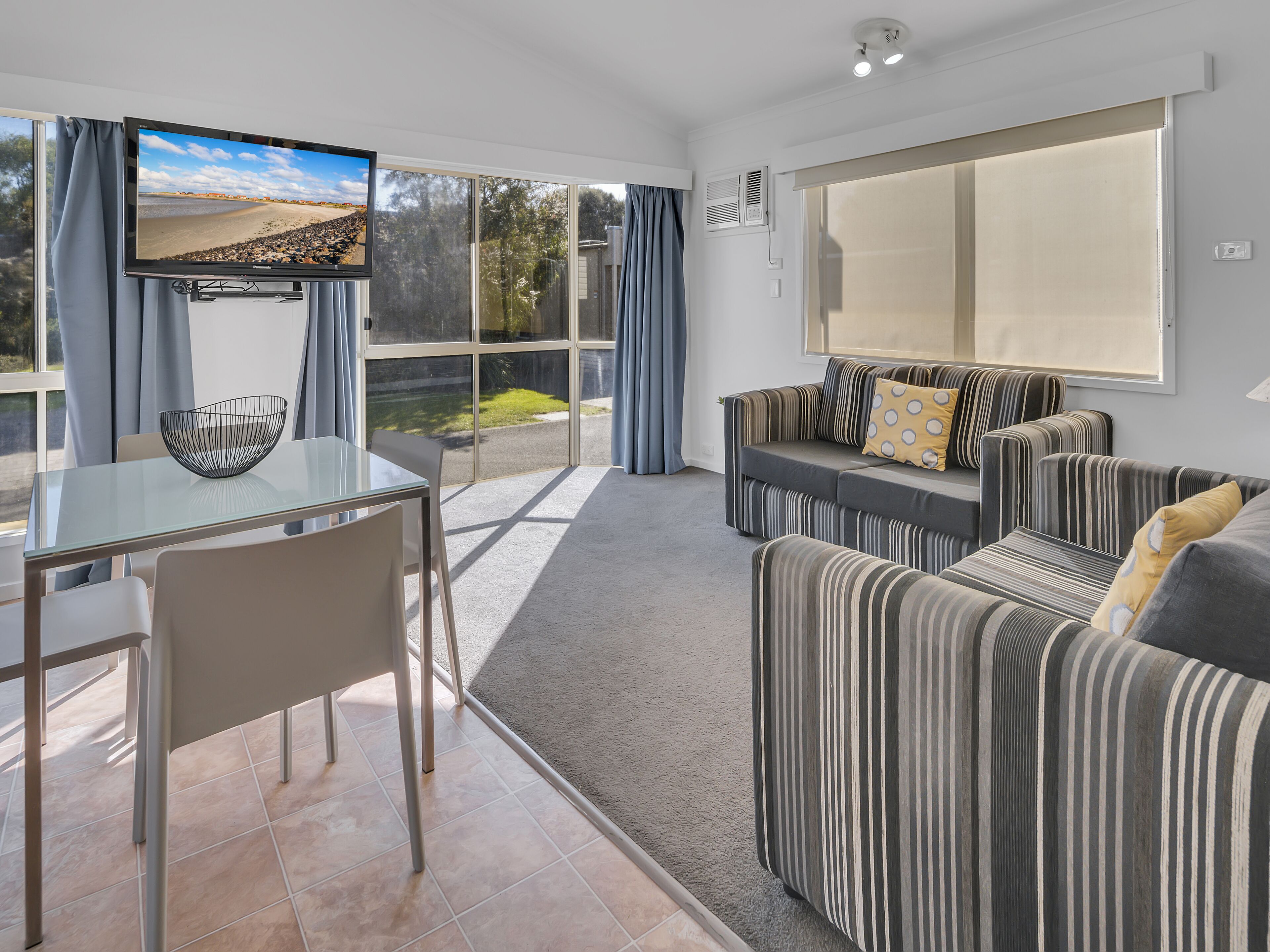 A modern living room with a glass dining table, striped sofas, and a television on the wall.