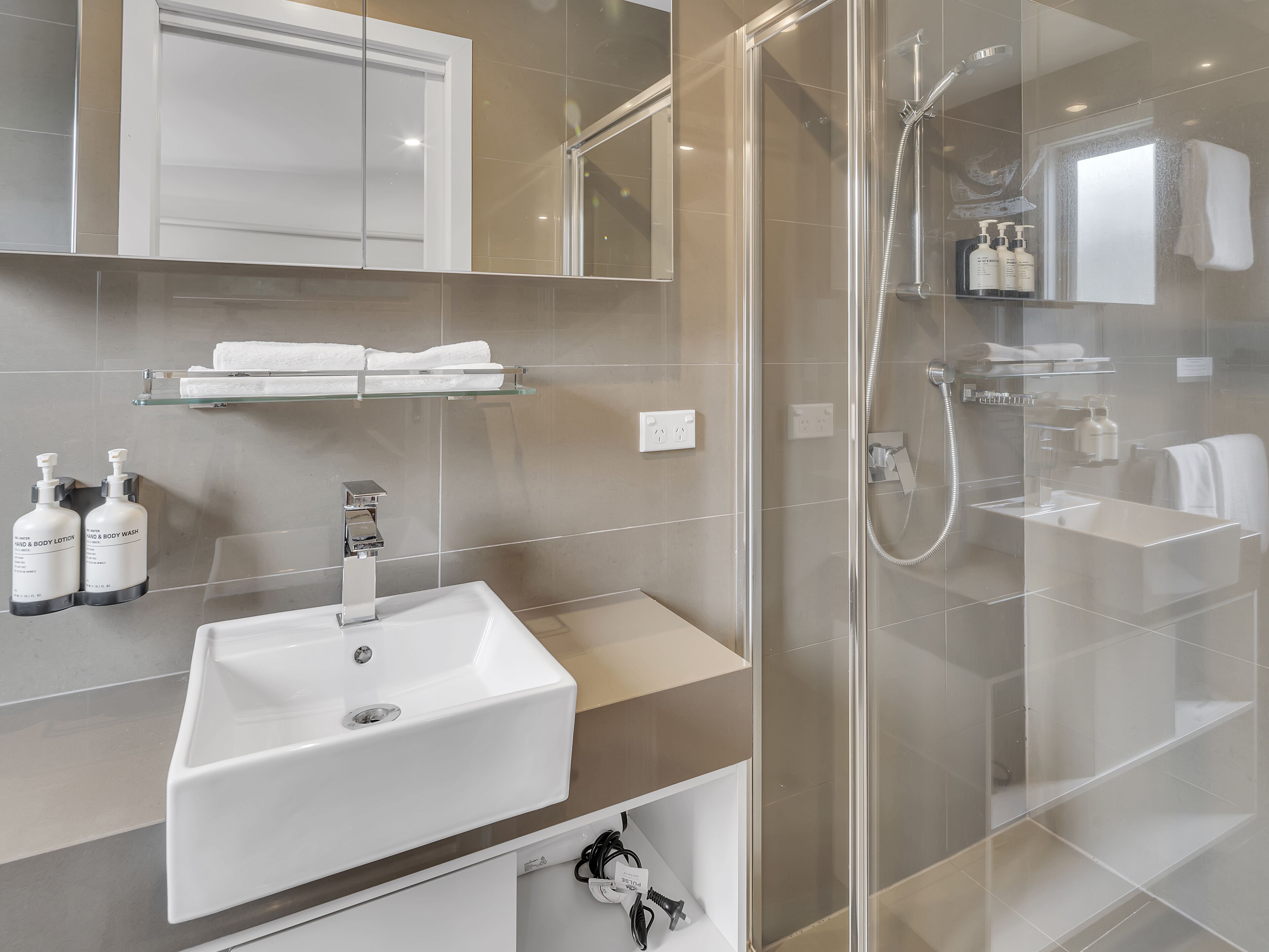 A modern bathroom with a glass-enclosed shower, a white sink, and a mirror above it. There are shelves with toiletries and a towel rack.