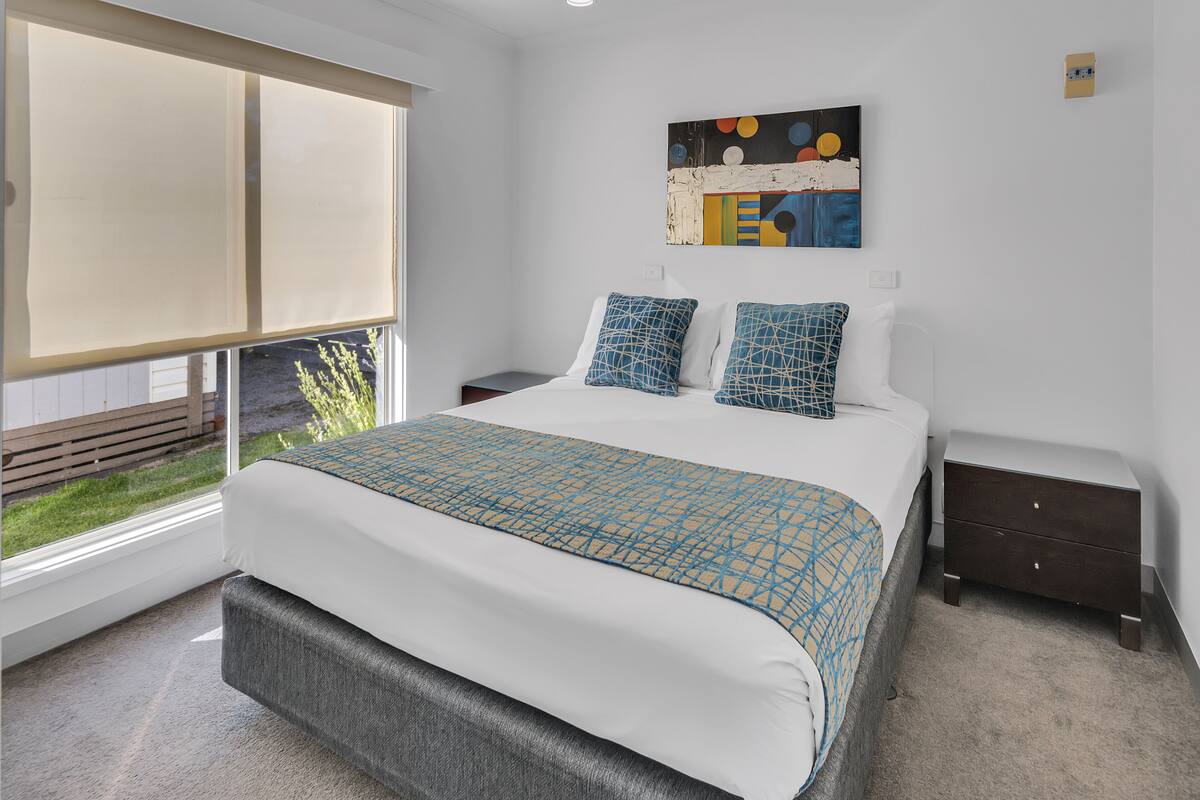 A modern bedroom with a large bed, a nightstand, a bedside table, and a window with a view of the outdoors.