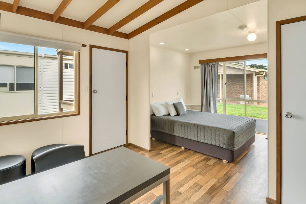 A bedroom with a bed, a desk, and chairs, a window, and a door leading to a balcony.