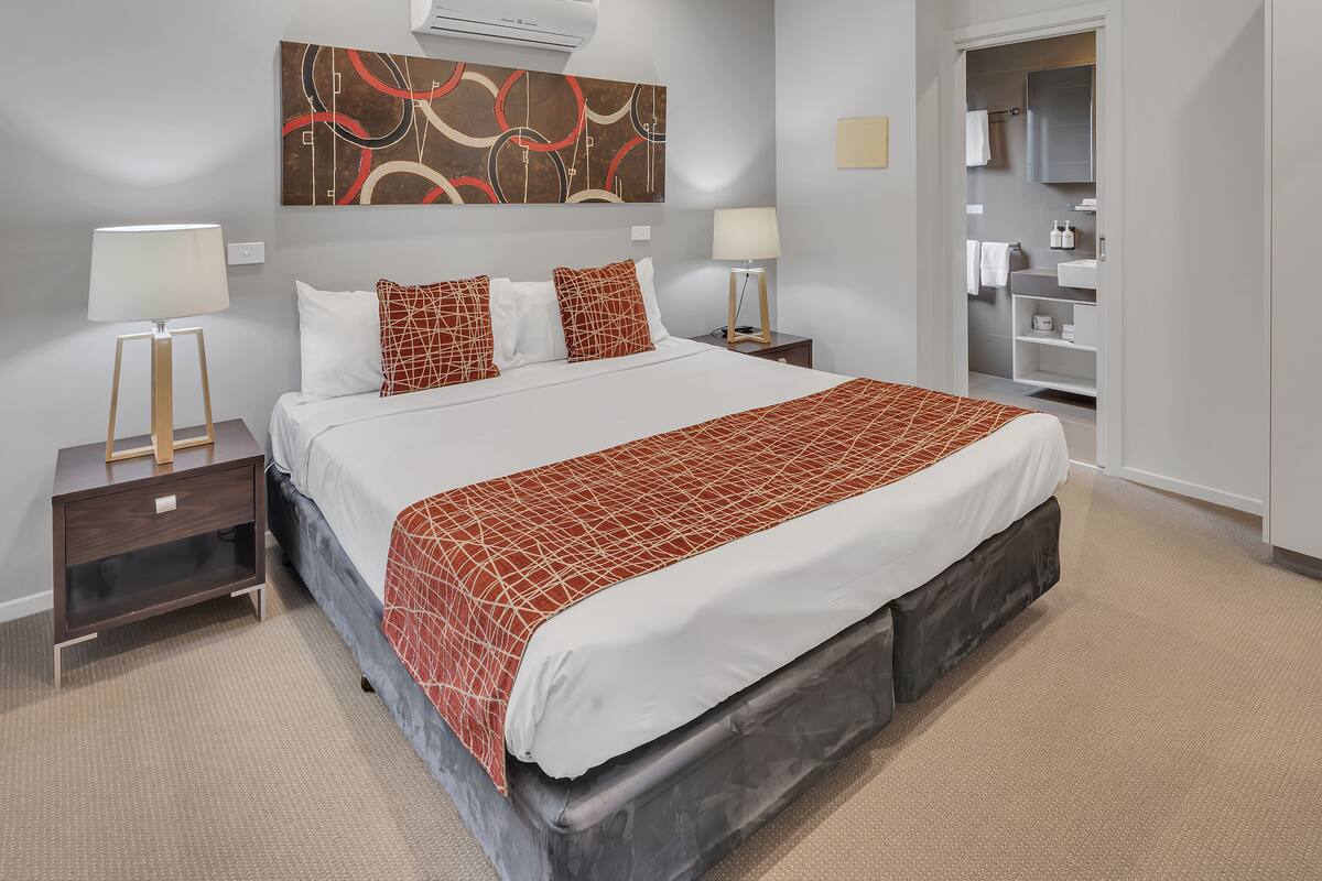 A modern hotel room with a large bed, two bedside tables with lamps, a wall-mounted air conditioner, and a bathroom visible through an open door.