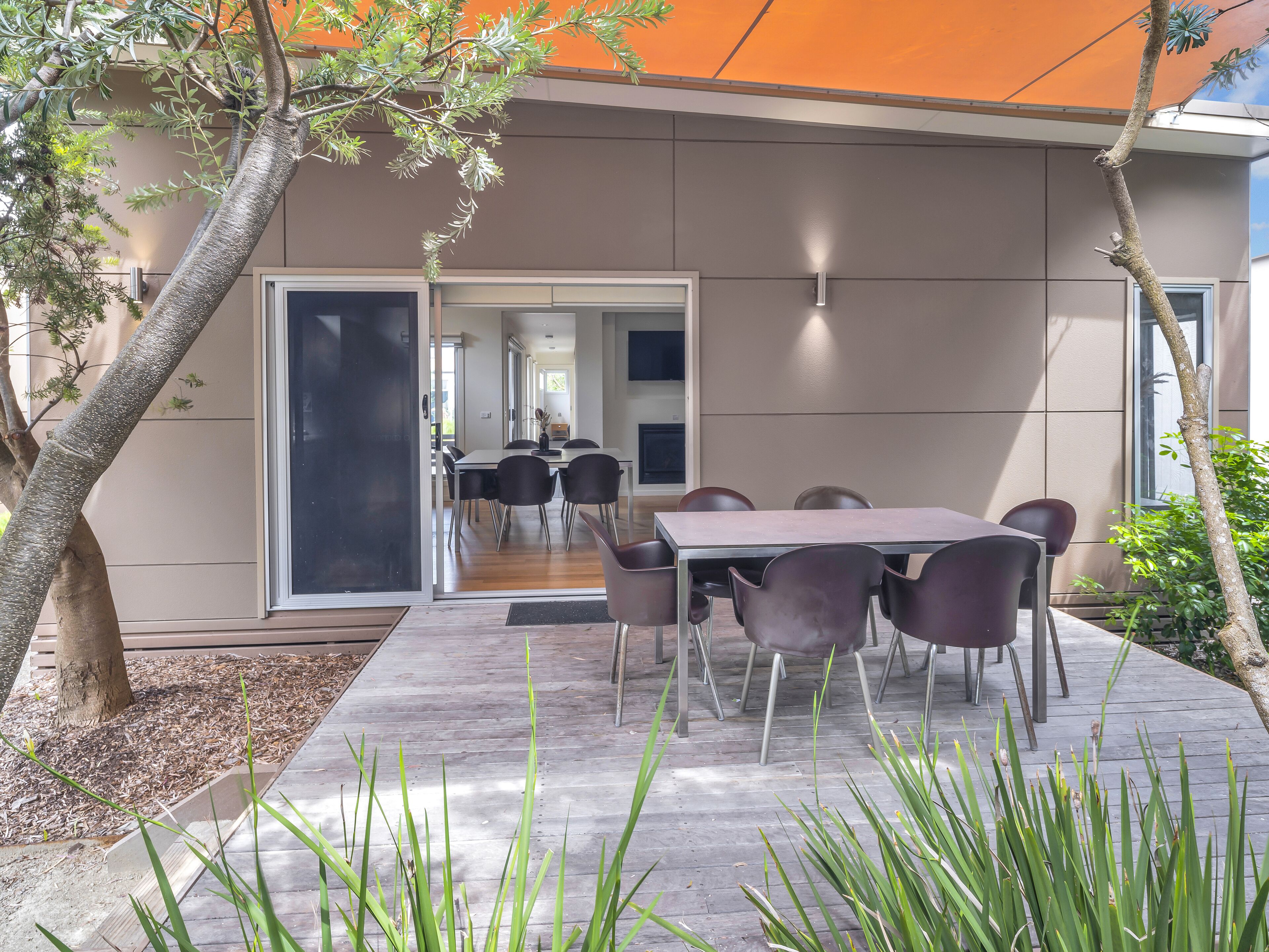 A modern outdoor dining area with a table and chairs, surrounded by plants and a tree.