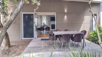 A modern outdoor dining area with a table and chairs, surrounded by plants and a tree.