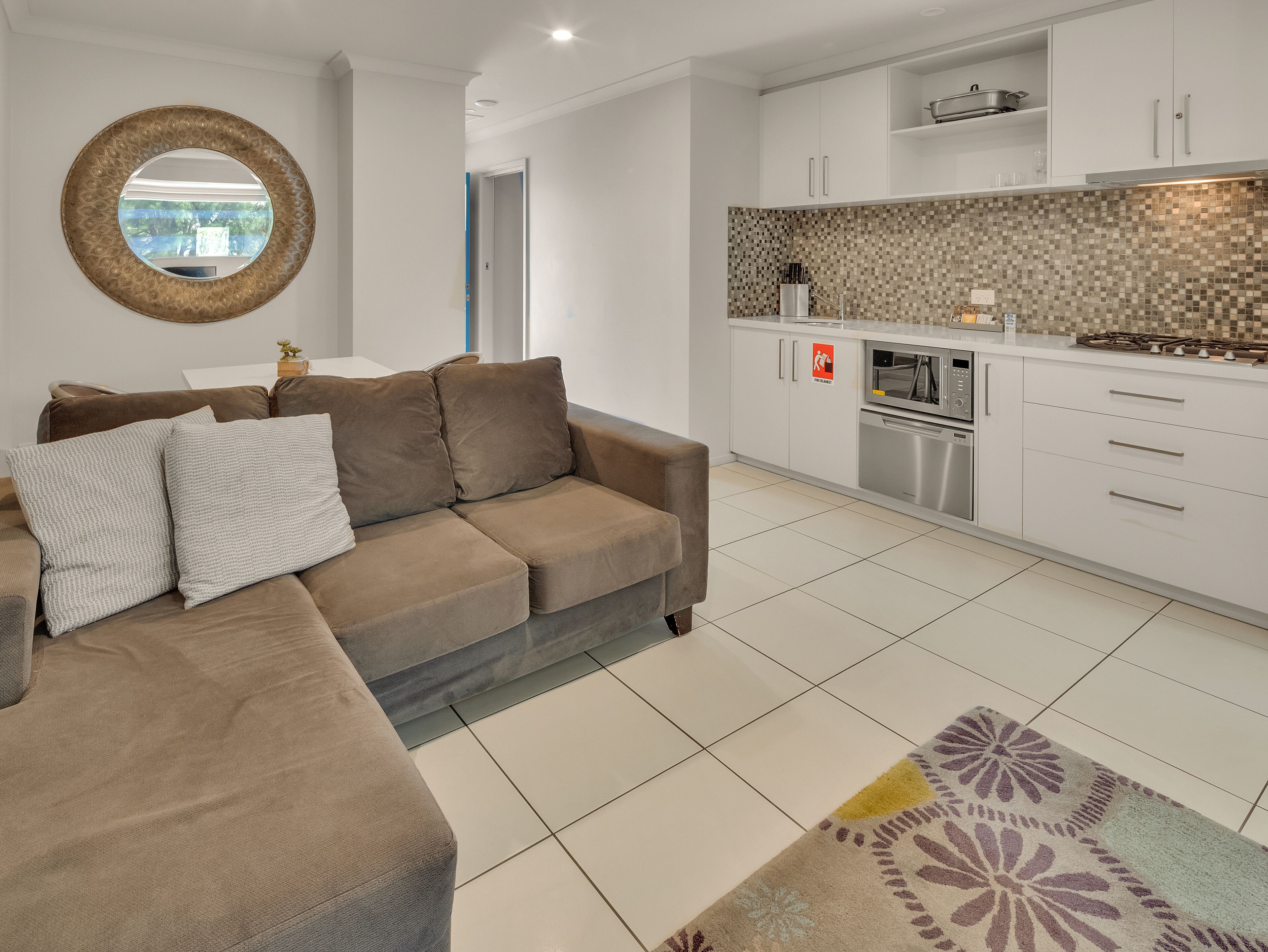 Un salon moderne avec un canapé d’angle marron, un tapis à motifs et une cuisine dotée de meubles blancs et d’un dosseret carrelé.