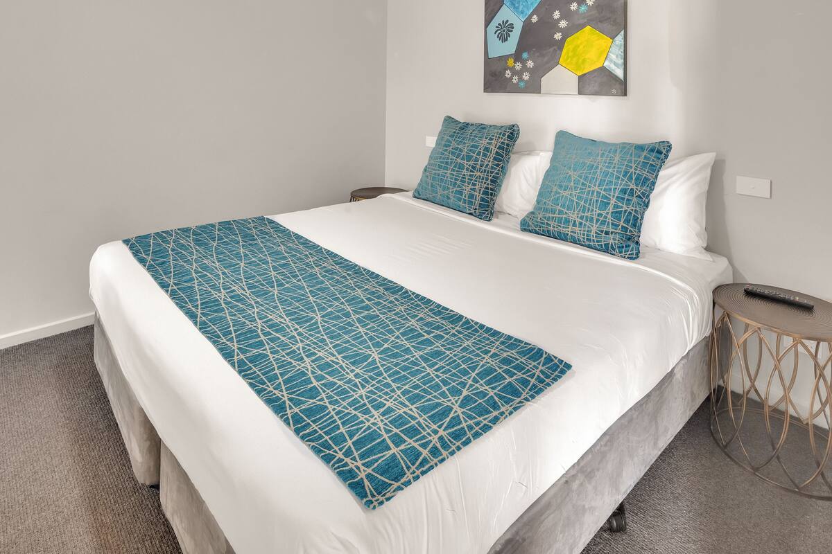 A neatly made bed with a geometric patterned bedspread and matching pillows, a small bedside table with a remote control, and a wall art piece above the bed.