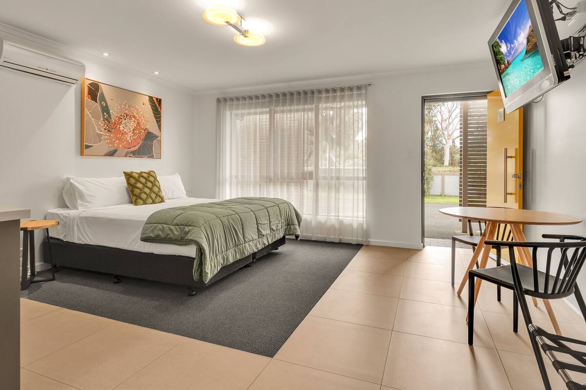 A modern hotel room with a bed, a TV, a dining table, and a window with sheer curtains.