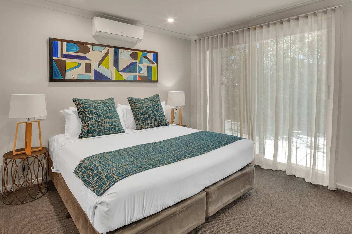 A modern hotel room with a large bed, two bedside tables with lamps, a geometric artwork on the wall, and sheer curtains on the window.