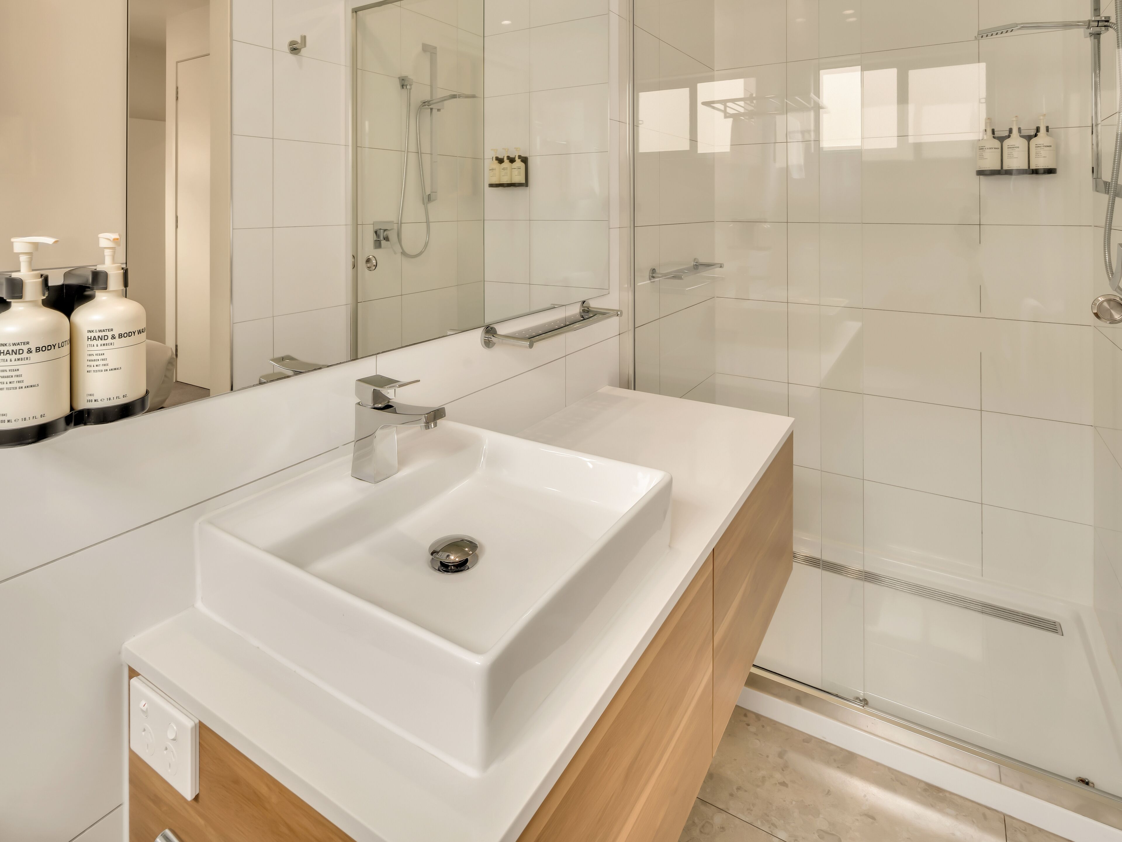 A modern bathroom with a white sink, a glass shower enclosure, and toiletries on a shelf.