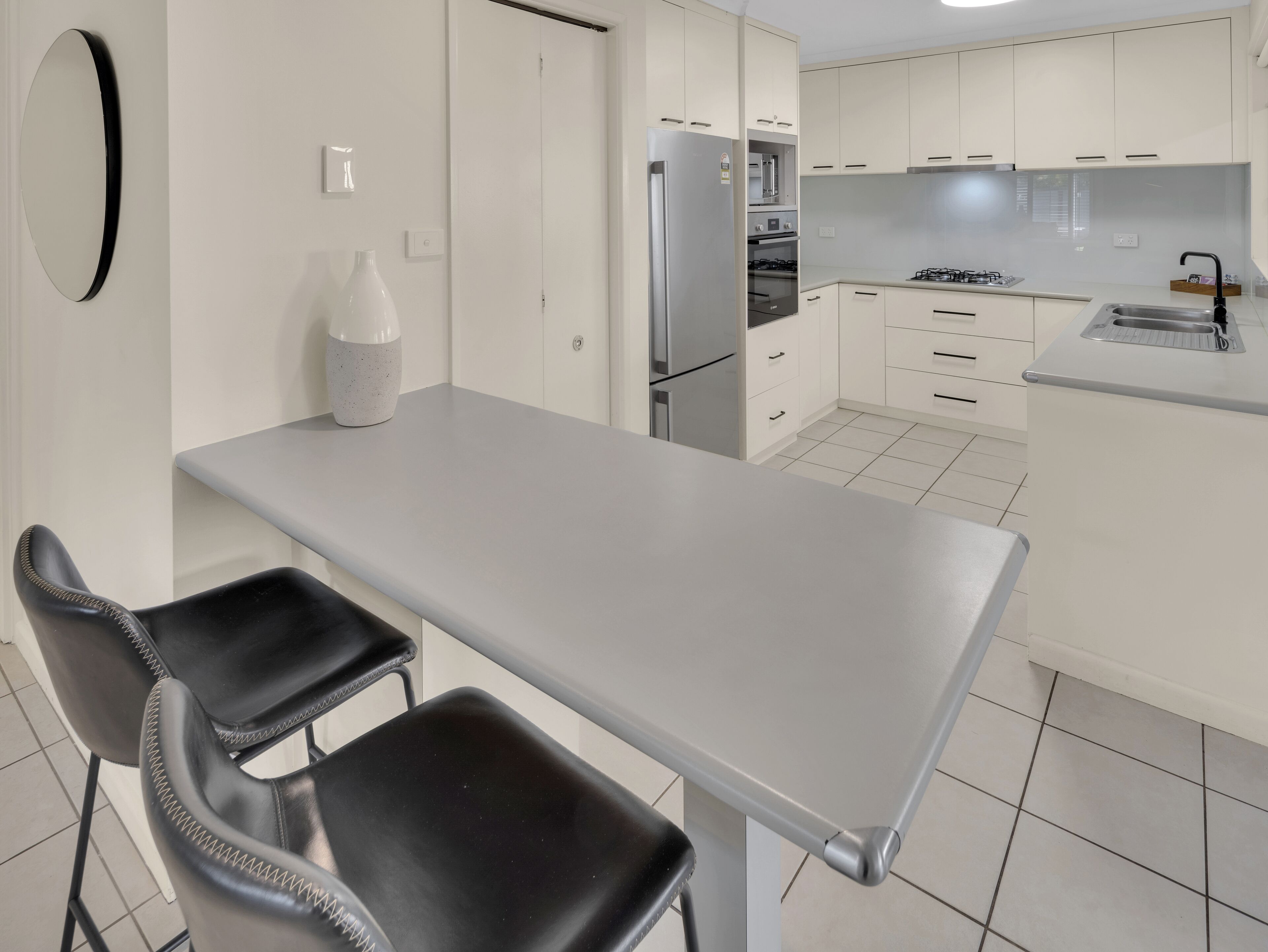 Una cocina moderna con armarios blancos, un refrigerador de acero inoxidable, una estufa a gas y una zona de comedor con una mesa y dos sillas.