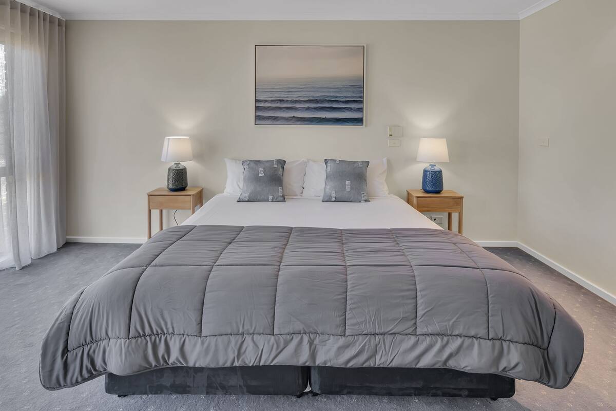 A hotel room with a large bed, two bedside tables with lamps, a painting of the ocean, and a view of the sea through the window.