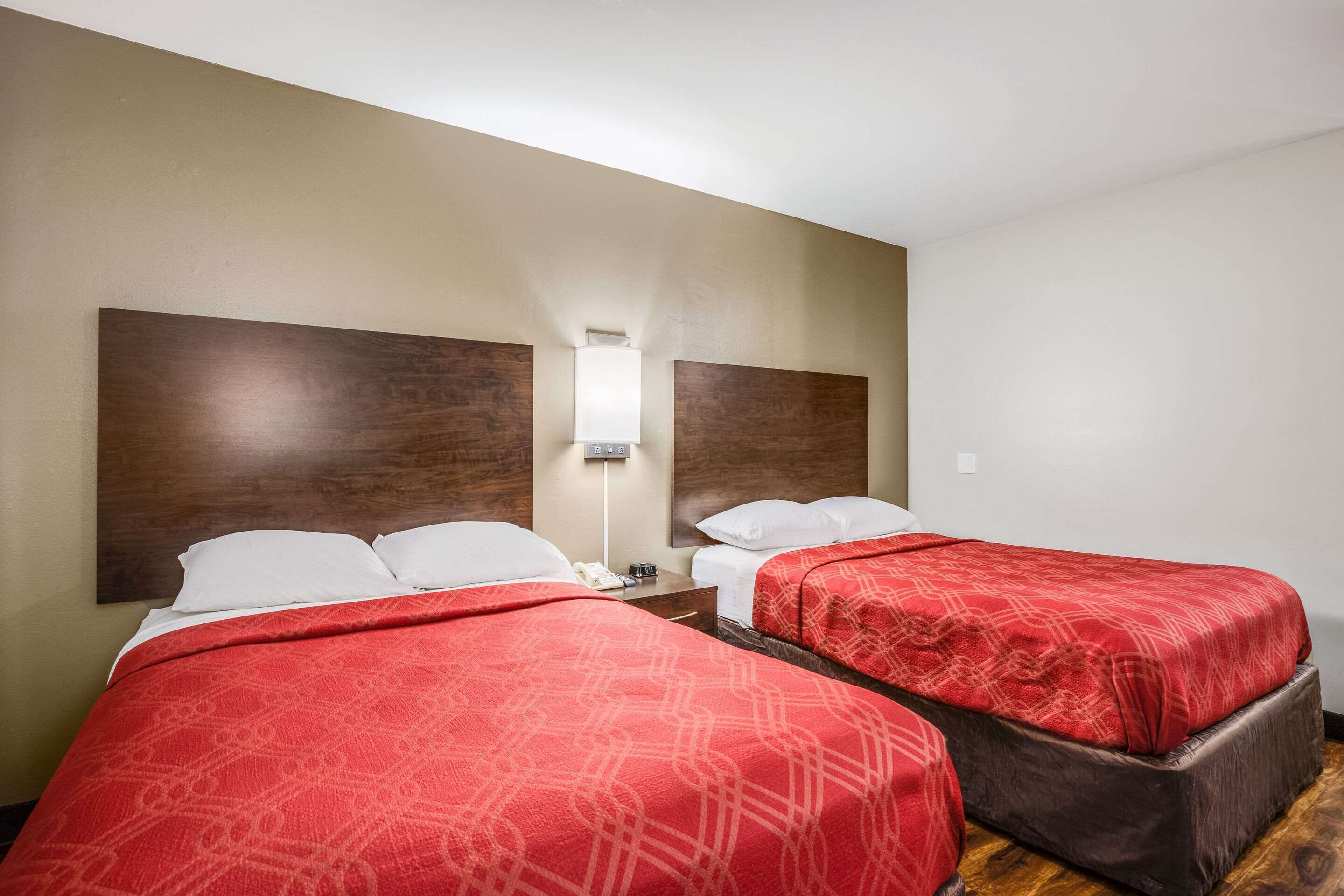 A hotel room with two beds, each with a red patterned bedspread, a nightstand with a lamp, and a wooden headboard.