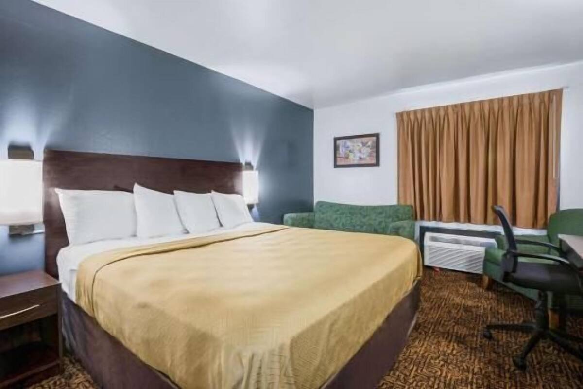 Habitación de hotel con cama, escritorio, silla, ventana con cortinas y una lámpara de pared.
