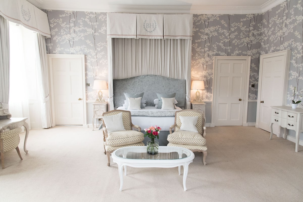 A bedroom with a large bed, two armchairs, a glass coffee table, and a dresser.