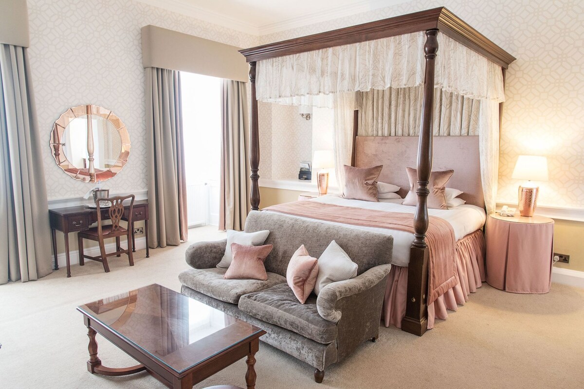 A four-poster bed, a desk with a chair, a small table, and a bedside lamp in a hotel room.
