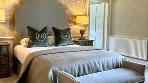 A bedroom with a large bed, two pillows with initials, a bench, a nightstand, a lamp, and a wall with a tree mural. - Balbirnie House (Glenrothes)