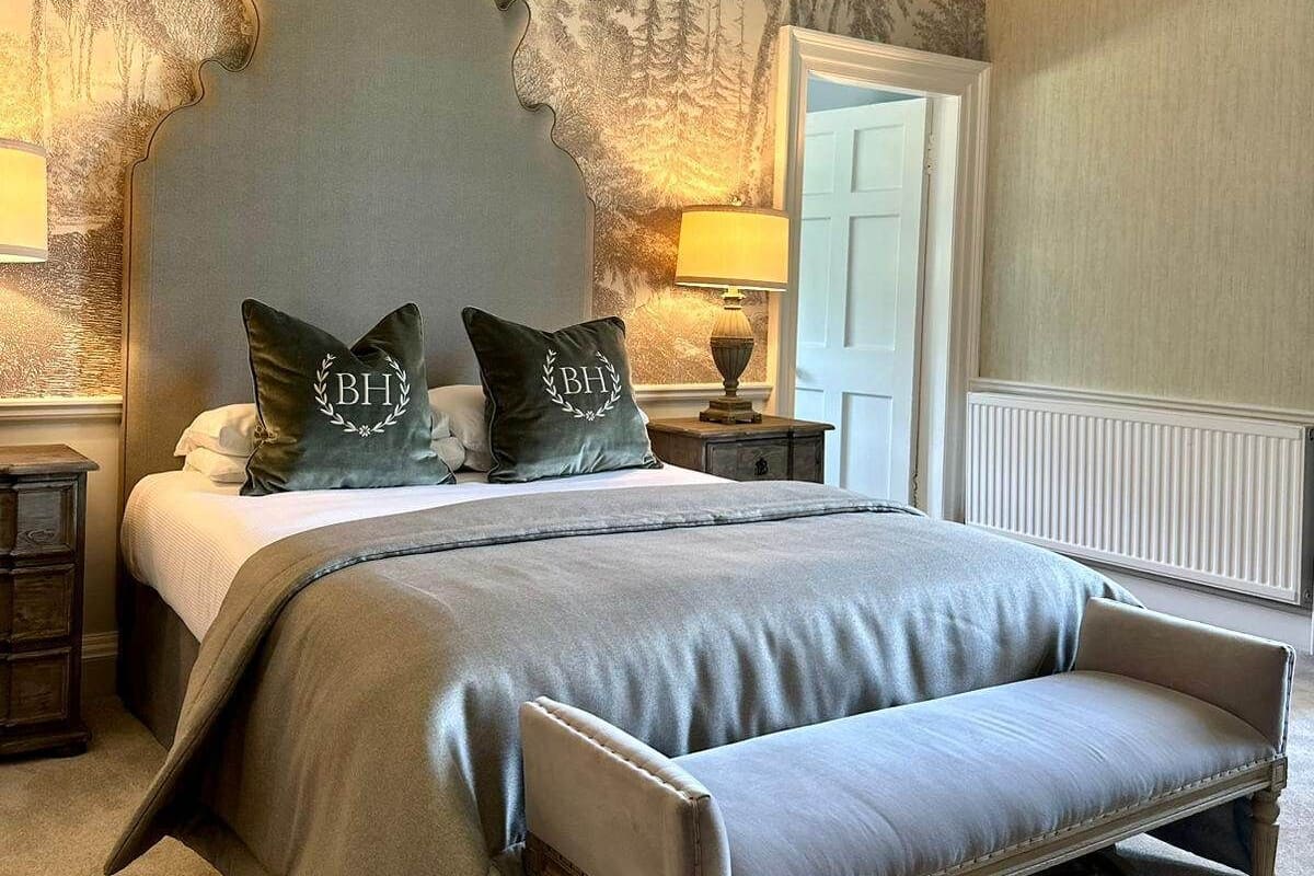 A bedroom with a large bed, two pillows with initials, a bench, a nightstand, a lamp, and a wall with a tree mural.