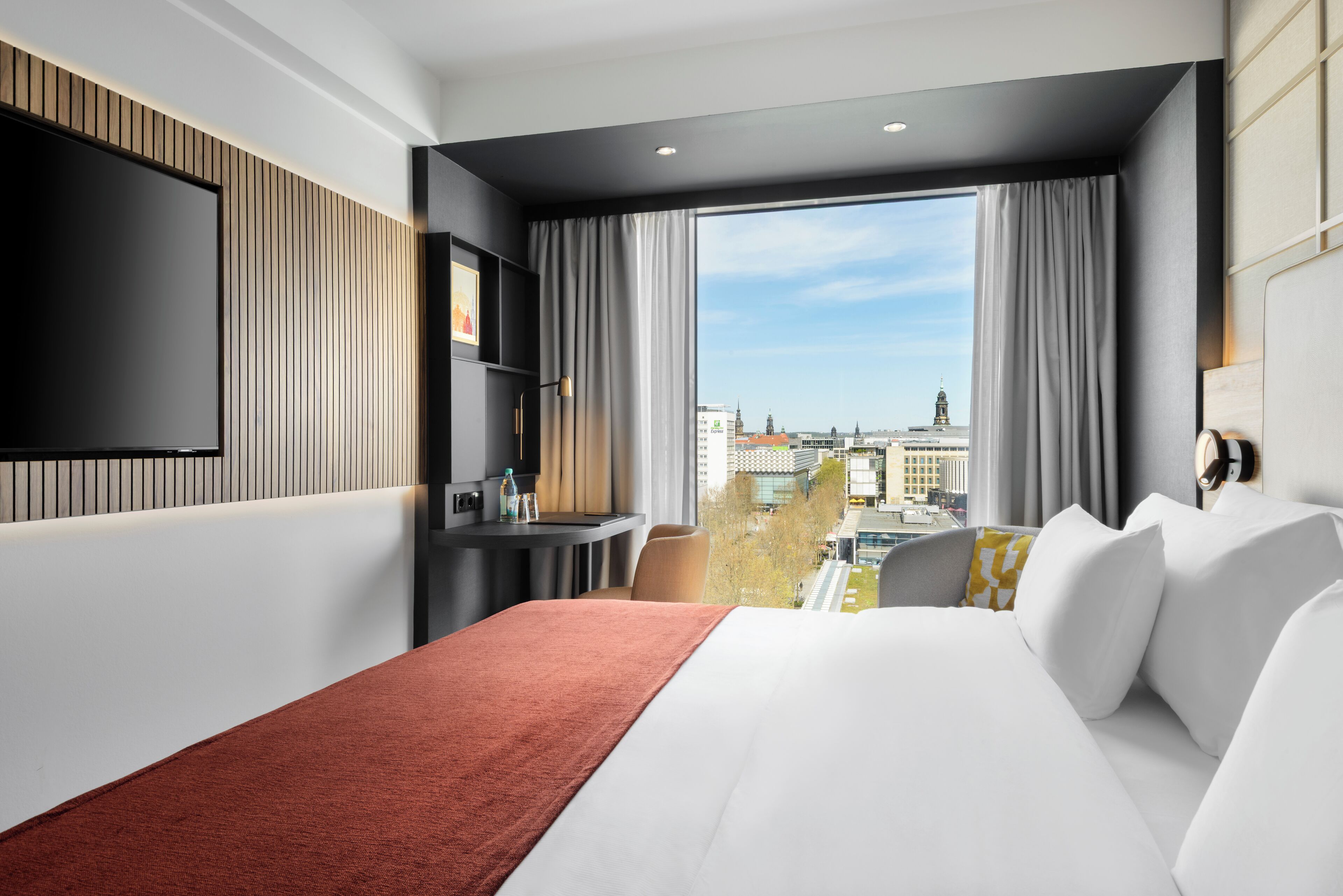 A hotel room with a large bed, a TV mounted on the wall, a small table, and a view of the cityscape.