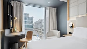Ett modernt hotellrum med en stor säng, ett skrivbord med en stol, ett litet bord och utsikt över staden genom stora fönster.