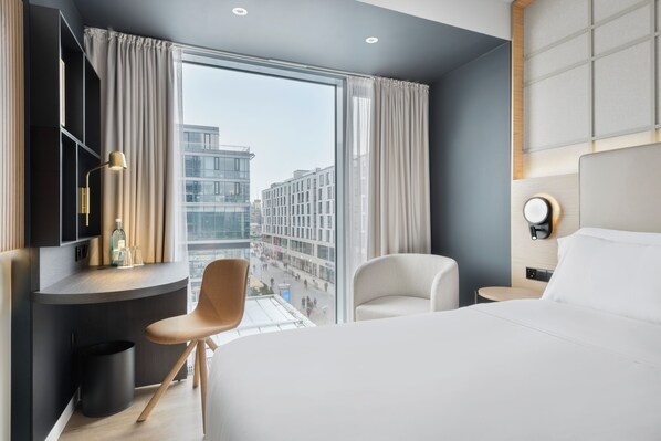 A modern hotel room with a large bed, a desk with a chair, a small table, and a view of the city through large windows. - Barceló Dresden Newa (Dresden)