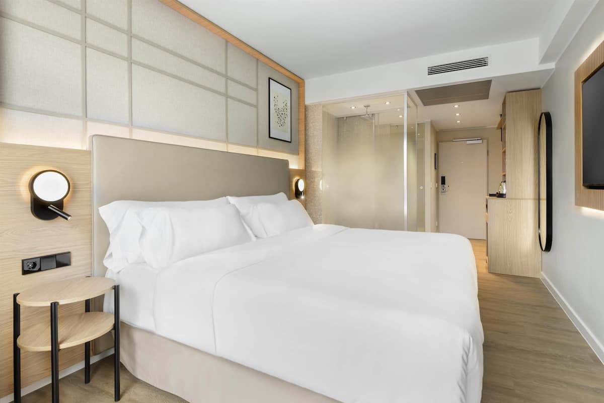 A modern hotel room with a large bed, bedside table, wall-mounted TV, and a bathroom visible through a glass partition.