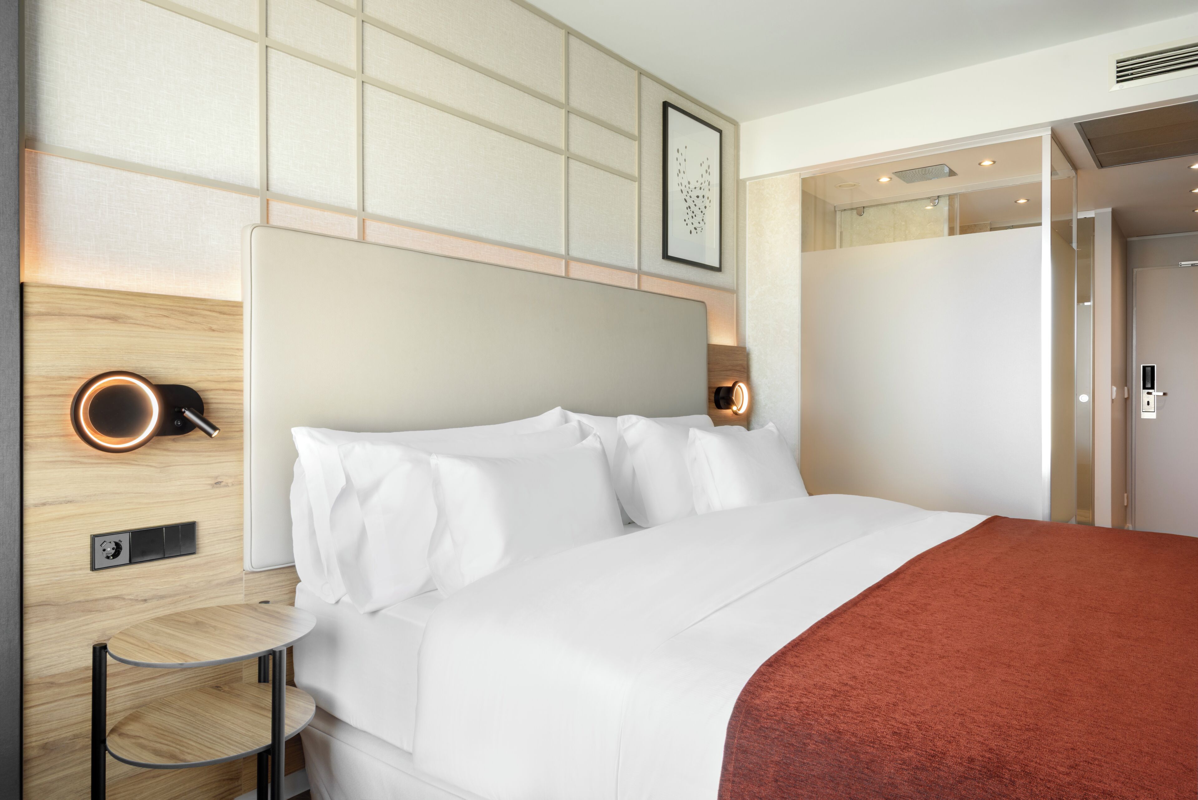 Una habitación de hotel con cama, mesita de noche, lámpara de pared y un cuadro enmarcado.
