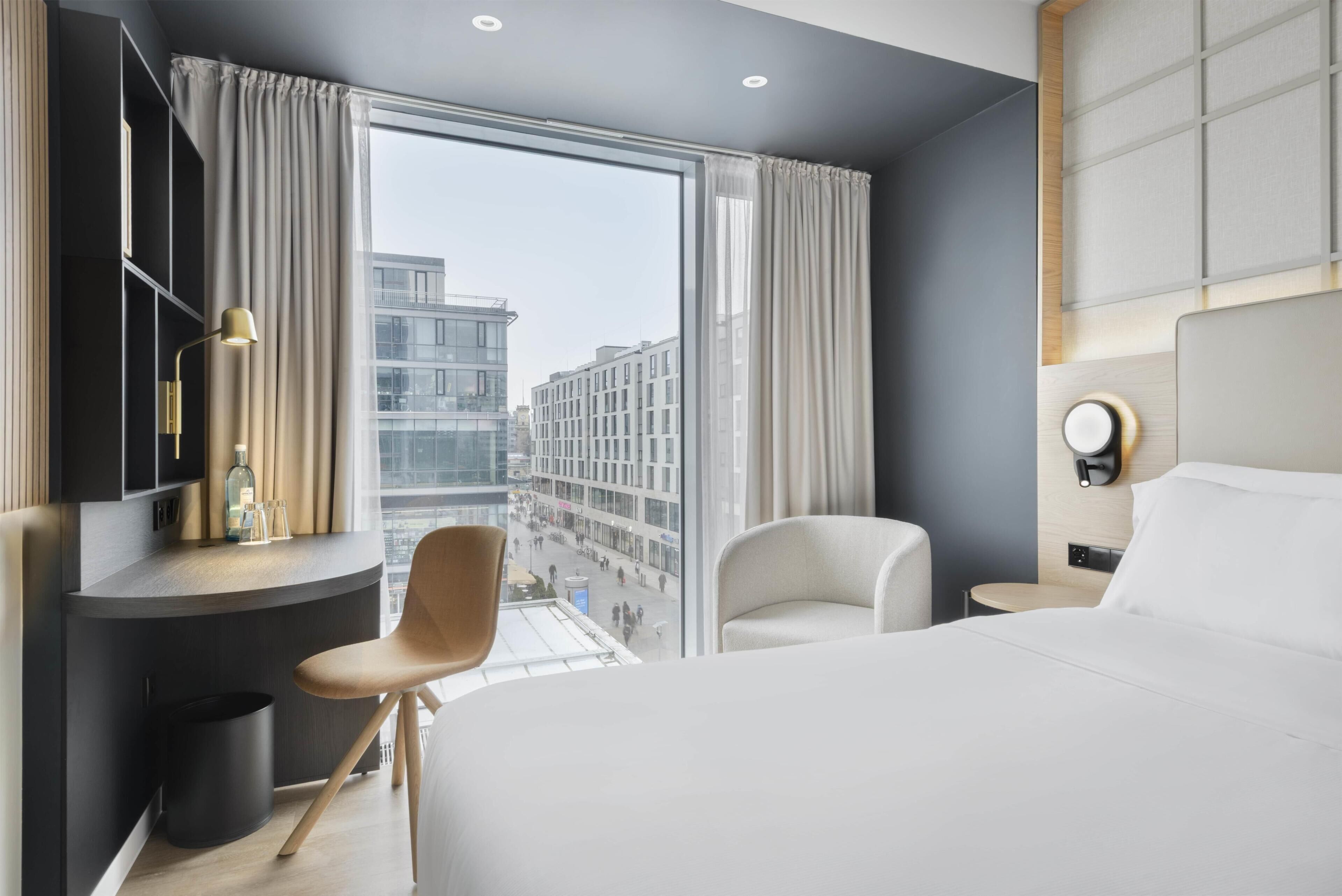 Quarto de hotel moderno com uma cama grande, uma mesa de escritório com cadeira, uma mesa redonda e vista para a paisagem urbana através de janelas amplas.