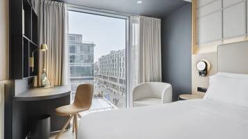 Quarto de hotel moderno com uma cama grande, uma mesa de escritório com cadeira, uma mesa redonda e vista para a paisagem urbana através de janelas amplas.