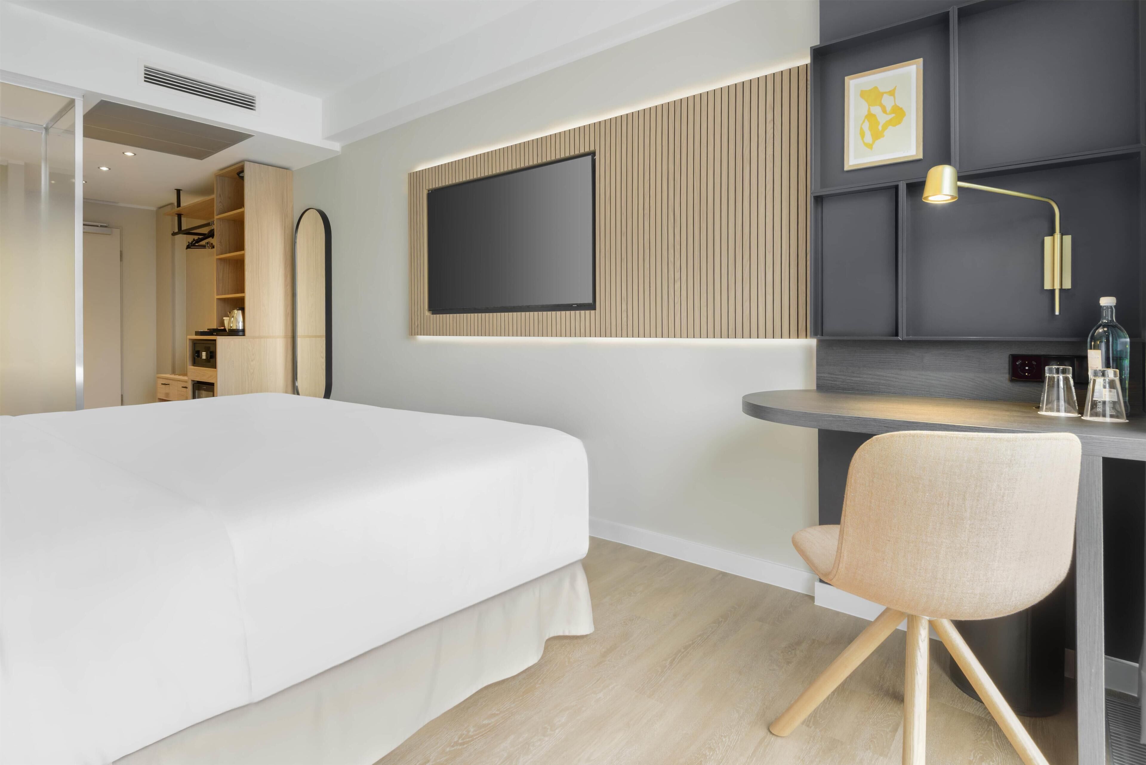 A modern hotel room with a bed, a desk, a chair, a TV mounted on the wall, and a small shelf with a lamp.