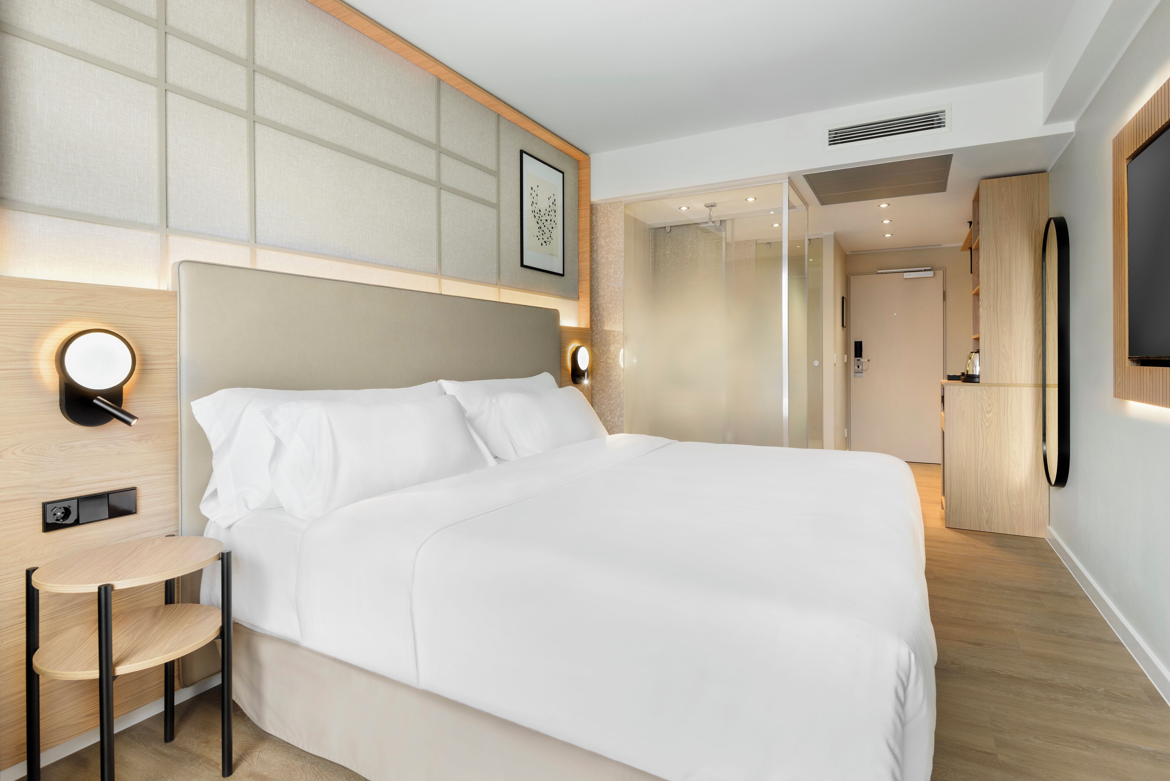Una habitación de hotel moderna con una cama grande, mesitas de noche, una pequeña zona de estar, ducha y un televisor montado en la pared.