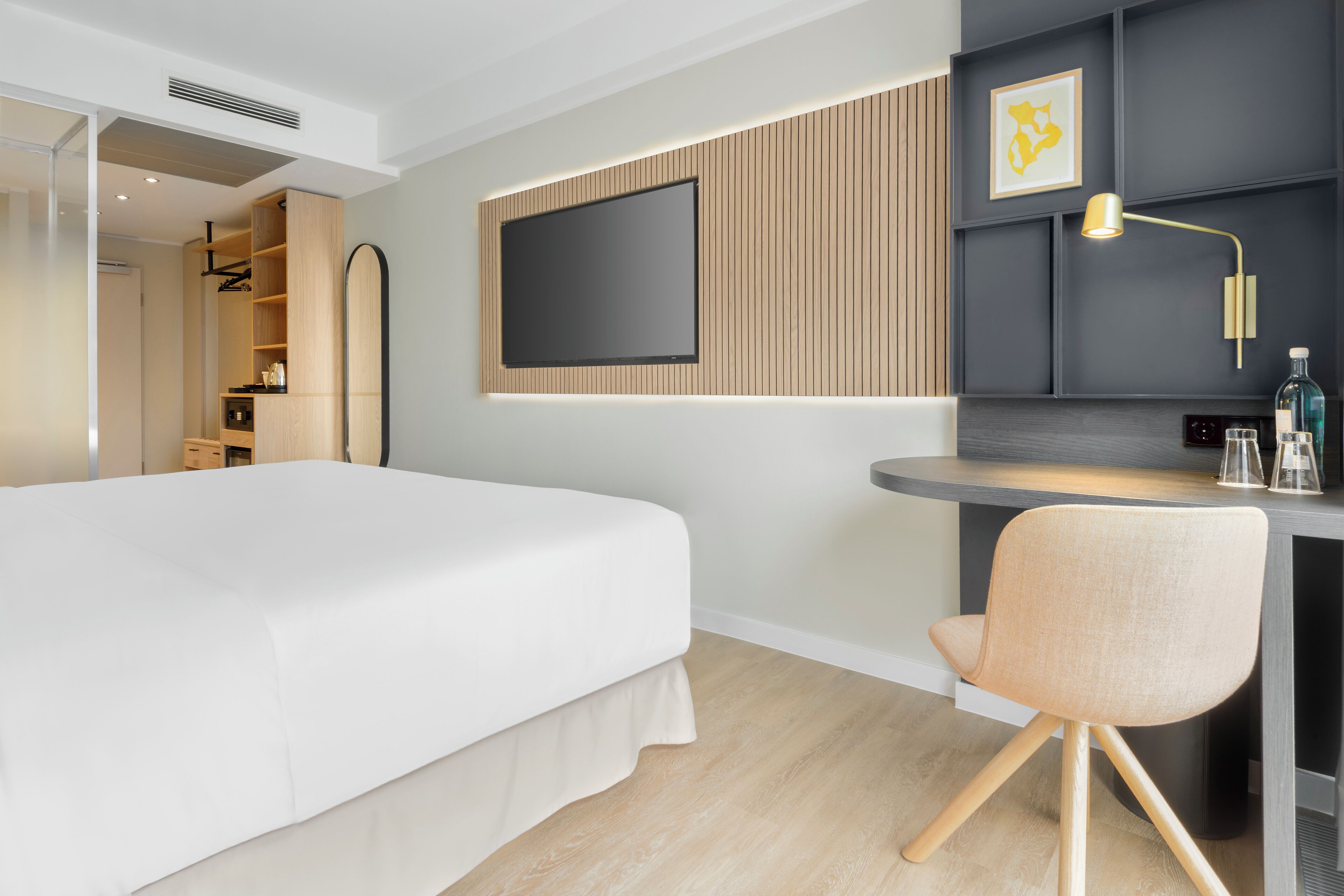 A modern hotel room with a large bed, a desk with a chair, a flat-screen TV mounted on the wall, and a small shelf with a lamp and decorative items.