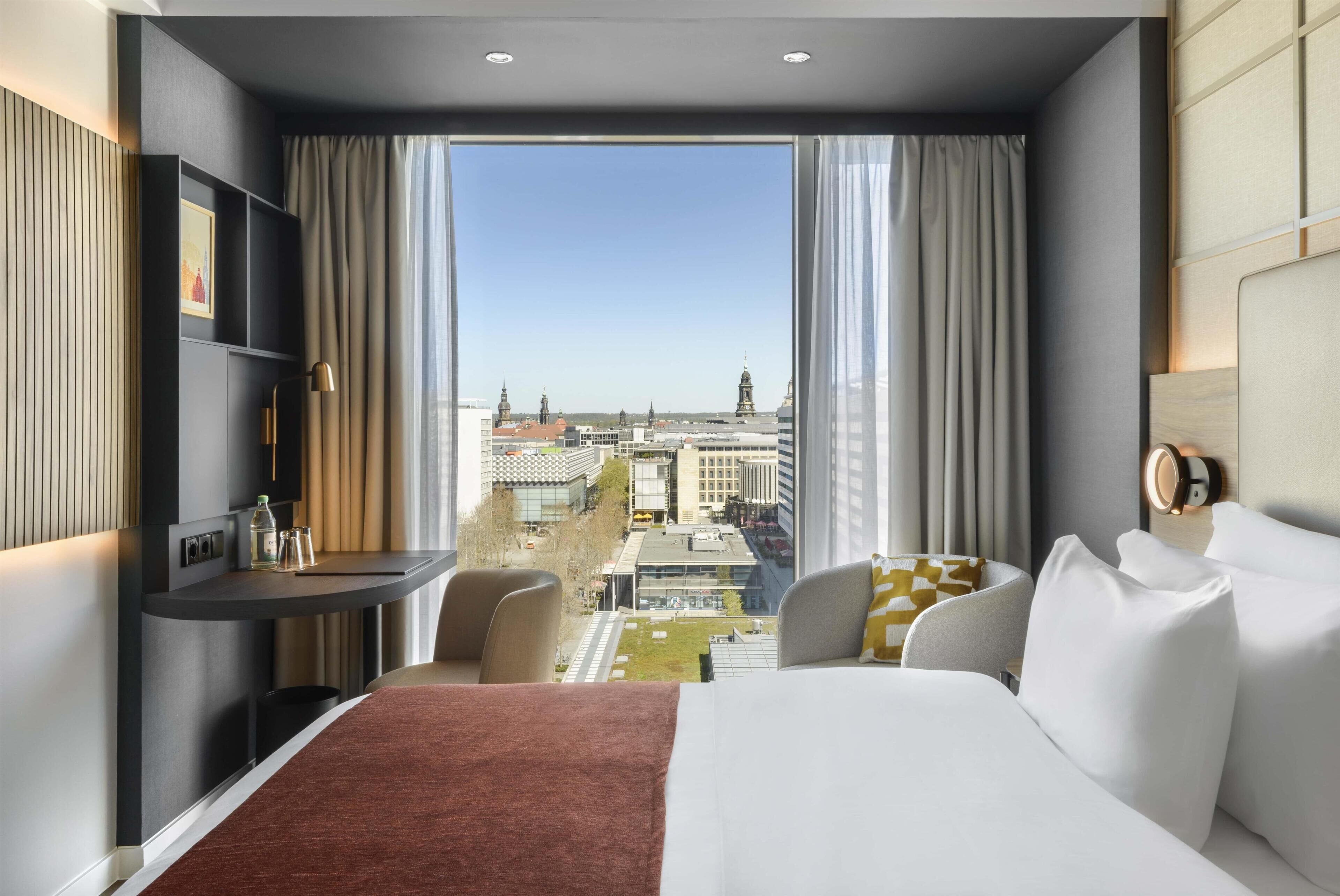 A hotel room with a large bed, a desk, a chair, and a view of the cityscape.