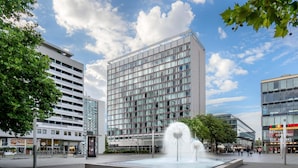 En modern byggnad med en glasfasad, en central fontän och ett omgivande, plattbelagt område.